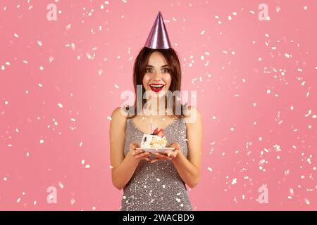 An excited woman with brown hair and red lipstick, wearing a glittering silver dress and pink party hat Stock Photo