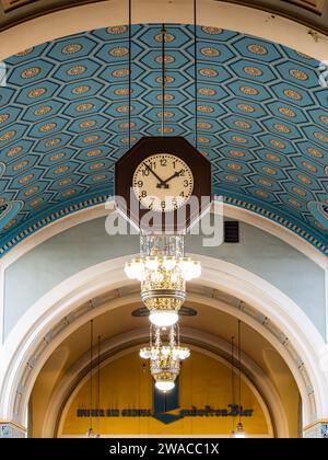 Clock in the train station hanging from the ceiling. Beautiful interior design of the ancient building with tiles on the wall and illuminating lights. Stock Photo