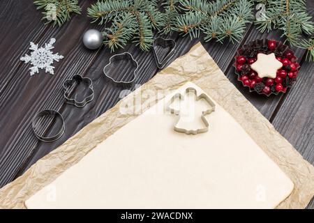 Dough for baking cookies on paper with cookie cutter molds and Christmas decorations. Metal form with berries and fir branches. Dark wooden background Stock Photo