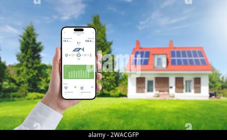 App on a mobile phone monitors the collection and consumption of electricity by solar panels. In the background is a house with solar panels on the ro Stock Photo
