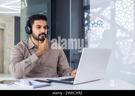 A serious thinking Hindus works inside an office with a laptop, a man in headphones listens to podcasts and audio books, a focused programmer encodes new software. Stock Photo