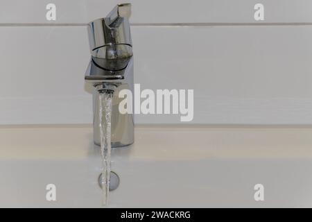 Close-up of water pouring from faucet into sink. White tiled wall and white ceramic sink. Background, copy space, text space Stock Photo
