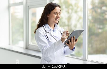 Cheerful young woman doctor standing by window, using digital tablet Stock Photo
