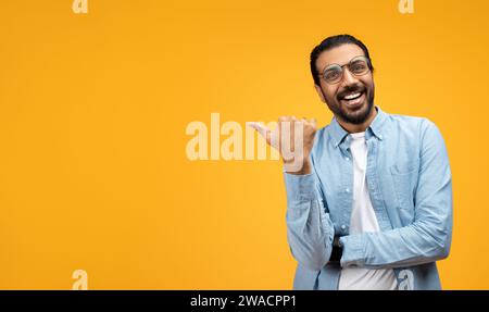 A joyful man with a beard and glasses points to his side with a thumb Stock Photo