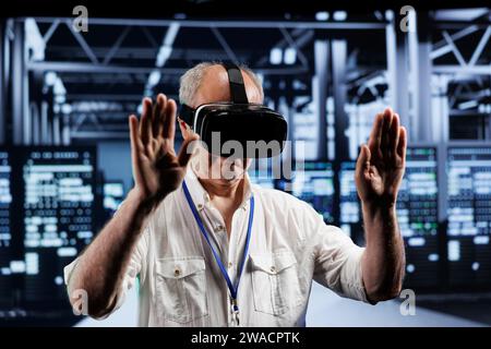 Elderly adept technician immersed in virtual reality at data center, doing infrastructure maintenance. Specialist using VR headset to optimize server clusters system performance Stock Photo