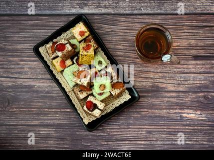 A rectangular black plate with a varied assortment of oriental sweets and a glass cup of hot tea with a spoon on a wooden table. Top view, flat lay. Stock Photo