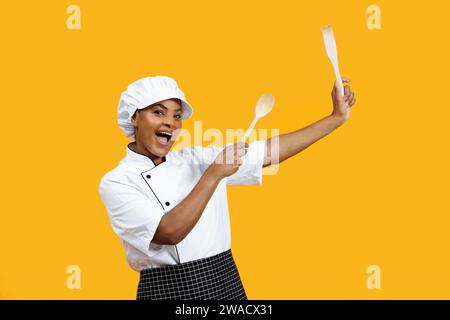 Cheerful young black female chef holding wooden spoons like drumsticks Stock Photo