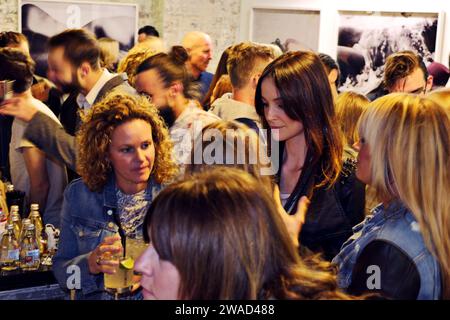 Crowd of people at an art opening in a Sydney warehouse art space, attractive women in the foreground, photographs on a wall in the background Stock Photo