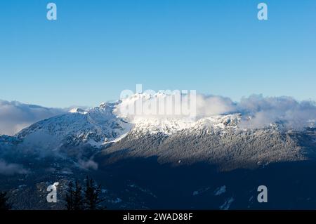 A dramatic view of snowcapped peaks engulfed in clouds, surrounded by thick forest, against a blue sky from Whistler Mountain, BC, Canada. Stock Photo