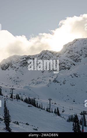 A dramatic view of a ski track and ski lifts against a snowy rocky slope and ridge with the sun partially visible through clouds in Whistler Mountain, Stock Photo