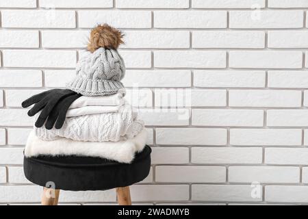 Stack of folded sweaters with hat and gloves on chair against white brick background Stock Photo