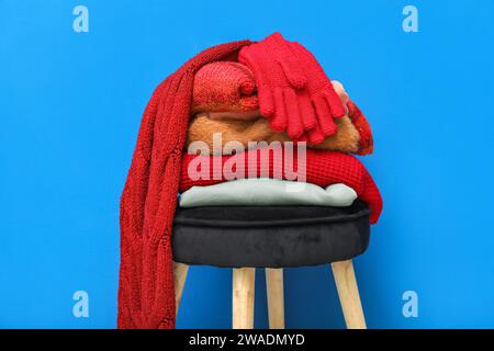 Stack of folded sweaters with scarf and gloves on chair against blue background Stock Photo