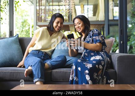Happy asian female friends talking and using smartphone at home. Domestic life, technology, friendship and lifestyle, unaltered. Stock Photo