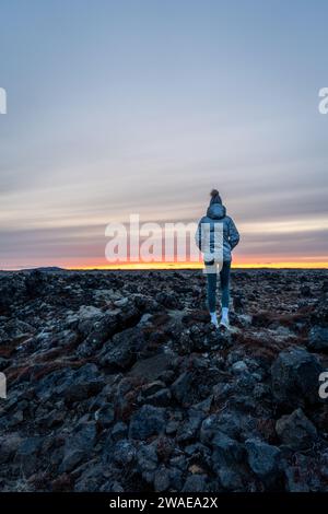 A silhouetted figure stands on a rocky outcrop in a grassy landscape, gazing out towards the horizon Stock Photo