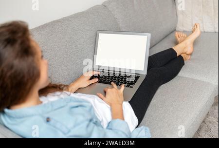 Over the shoulder view laptop with blank screen on laps of woman. Woman uses her laptop while relaxing at home on sofa. Stock Photo