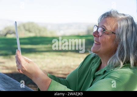 mature white-haired woman smiling looking at her digital tablet on a video call, in the background a green grassy meadow Stock Photo