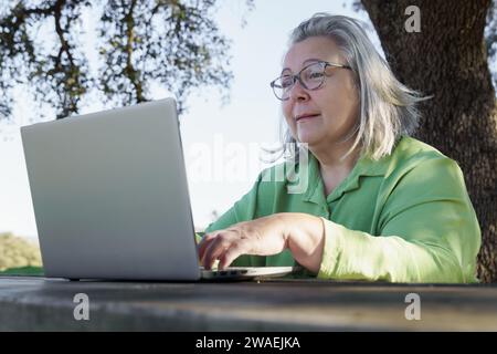mature woman with white hair and glasses smiling as she works at her computer on a wooden table in the countryside. Stock Photo