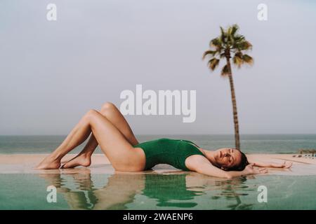 A young woman in a green swimsuit posing on a poolside with a palm tree in the background Stock Photo