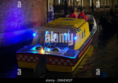Three paramedics on an ambulance boat driving with flashing blue lights to a night-time rescue mission in a narrow canal in Venice. Stock Photo