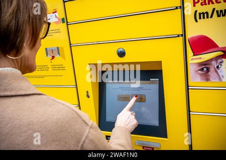 Innovative Paketlogistik - Nutzung der DHL Packstation 04.01.2024, Bad Camberg: Symbolfoto, Illustrationsbild, Symbolbild, Illustrationsfoto Das Bild zeigt eine Frau im Anschnitt, die mit ihrem Smartphone über den Touchscreen einer DHL Paketstation interagiert, um ein Paket einzureichen. Sie ist konzentriert auf die digitale Schnittstelle fokussiert, während ihre Hand das Mobiltelefon hält und die entsprechenden Eingaben auf dem Bildschirm der Paketstation tätigt um das Paket einzulegen. Die Packstation, ein Paketautomatensystem von DHL, bietet seit 2001 in über 12.500 Standorten deutschlandwe Stock Photo