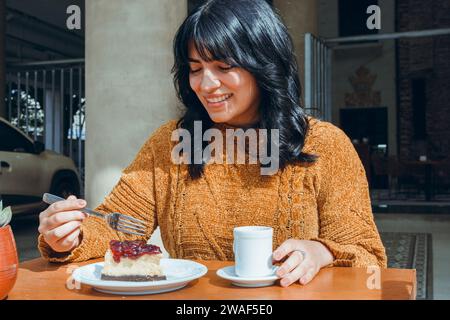 young latin woman with curlers, sitting outdoors happy smiling eating cutting slice of cheesecake with fork and drinking coffee, copy space. Stock Photo