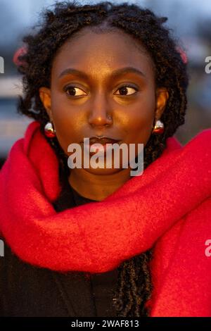 A portrait of a young Black woman captured during the golden hour, which casts a warm glow on her features. She is wrapped in a bright red scarf that contrasts with her dark skin, giving the image a vibrant look. Her gaze is directed away from the camera, implying thoughtfulness. The image has a shallow depth of field, with the focus on her eyes, enhancing her reflective expression. The lighting and color contrast make the image visually appealing. Golden Hour Contemplation. High quality photo Stock Photo
