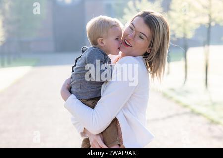 A radiant scene unfolds as a young Caucasian woman lifts a toddler high in a loving embrace, his small arms wrapped around her neck. The child's gesture, a sweet kiss on the cheek, brings a smile of pure joy to the woman's face. The soft morning light enhances the warmth of the moment, highlighting the bond of affection that connects them. The park's backdrop offers a calm and inviting space for such delightful interactions. Joyful Embrace in the Morning Sun. High quality photo Stock Photo