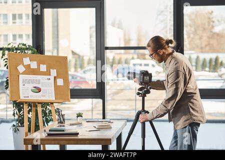 entrepreneur in casual attire adjusting digital camera near desk and corkboard with charts in office Stock Photo