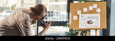 office manager in casual clothes adjusting digital camera near corkboard with infographics, banner Stock Photo