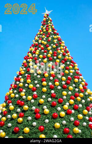 christmas tree with decorates balls and large beautiful star at the top with 2024 Happy New Year made from chain links on clear blue sky Stock Photo