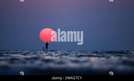 Silhouette of a man on the paddleboard against the setting sun. Paddleboard in the sea at sunset, side view . Young man on paddle boarding during a Stock Photo