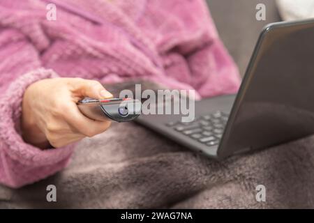 A woman sits on the sofa at home with a TV remote control in her hand and a laptop on her lap. Stock Photo
