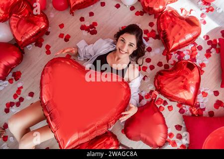 Top view of young woman lying on wooden floor holding a big red heart shaped balloons in Valentines Day. Girl is smiling, wearing casual outfit. Floor Stock Photo