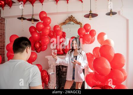 Joyous moment between young couple celebrating with toast Valentines day near red balloons, white fireplace. Woman laughing, holding glass, about to c Stock Photo
