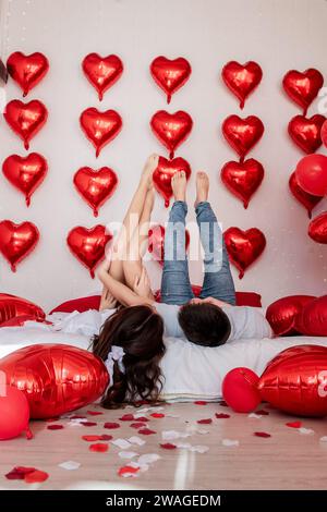 Crazy couple having fun, lying, legs up on white bed near red heart shaped balloons, rose petals, pillows. Man and woman are fooling around, jumping. Stock Photo