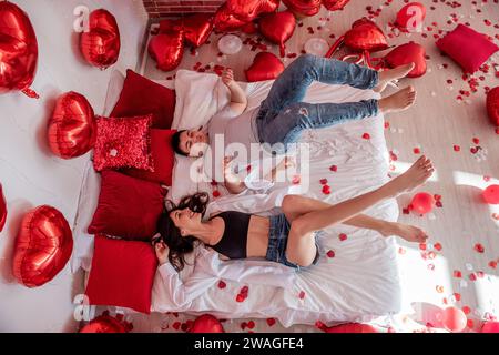 Top view of crazy couple having fun, lying on white bed near red heart shaped balloons, rose petals, pillows. Man and woman are fooling around, jumpin Stock Photo