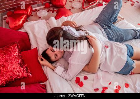 Couple in love hugs tightly on white bed among red balloons, pillows, hearts, rose petals. Man and woman look at each other, spend Valentines Day home Stock Photo