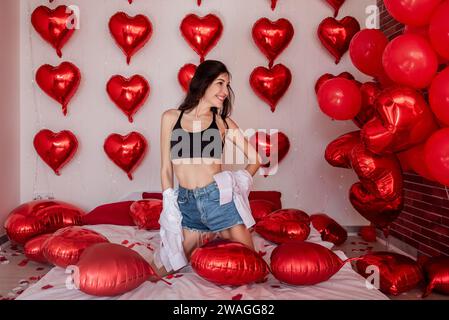 Young woman kneels on bed with rose petals, among inflatable red heart-shaped balloons. Girl blogger poses in white shirt short denim shorts in decora Stock Photo