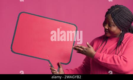 African American beauty holding a red speech bubble mockup for text messages and notes in her right hand. Black lady nodding in agreement with the content of the word-bubble copyspace template. Stock Photo