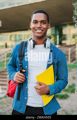 Vertical individual portrait of a african american student young man carrying a backpack and note books at university campus. Teenage male smiling and looking at camera. High school guy staring front. High quality photo Stock Photo