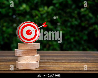 Big 3d target icon on the top of round wooden blocks stack on green natural leaves background with copy space. Business goal and success, effort and d Stock Photo