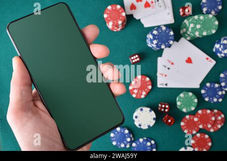 Concept of playing online casino games with hand holding mobile device and background objects on a green felt mat. Top view. Stock Photo