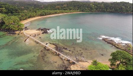 A picturesque wooden bridge spans across an open space leading to a pristine beach surrounded by lush tropical jungle Stock Photo