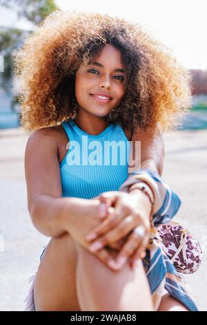 Happy young black female in casual crop top with curly Afro hairstyle smiling and looking at camera while sitting with hands on raised knee over sport Stock Photo