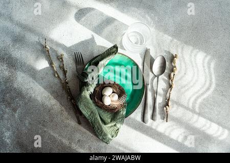 Top view of Easter table setting, showcasing a vibrant green ceramic plate with a small nest with speckled eggs and feathers, placed on gray surface w Stock Photo