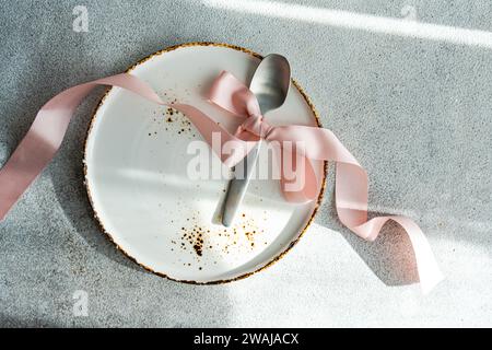An empty white plate with golden rim, adorned with a soft pink ribbon and a silver spoon, casting elegant shadows on a textured surface Stock Photo