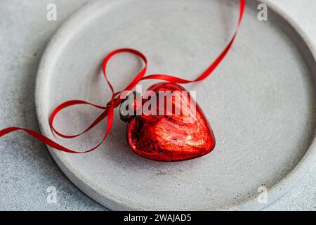 Closeup of simple yet evocative composition, with a concrete gray round plate with a striking red heart-shaped ornament, wrapped in a vibrant red ribb Stock Photo