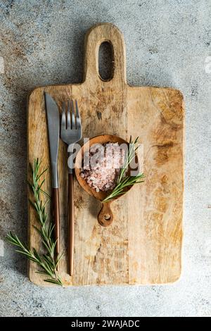 From above rustic kitchen setup on wooden cutting board with a fork, knife, a sprinkle of pink Himalayan salt, fresh rosemary sprigs, laid out on a te Stock Photo
