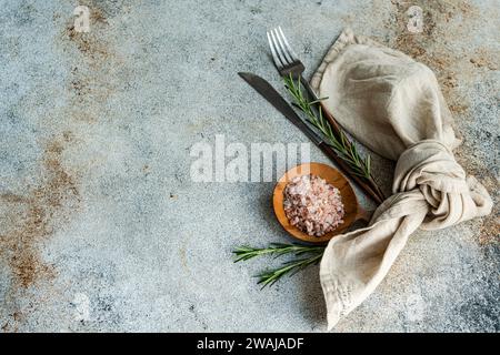 From above wooden spoon with pink Himalayan salt, surrounded by rosemary sprigs and a folded linen napkin alongside a knife and fork, all arranged nea Stock Photo