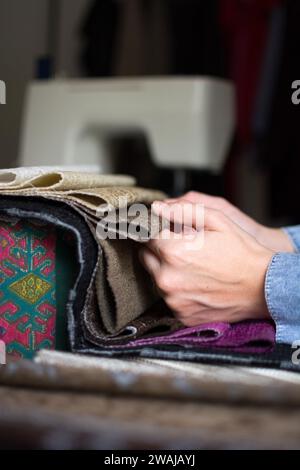 A close-up of a person's hand rifling through various fabrics, with a sewing machine in the background, getting ready for a sewing project. Stock Photo